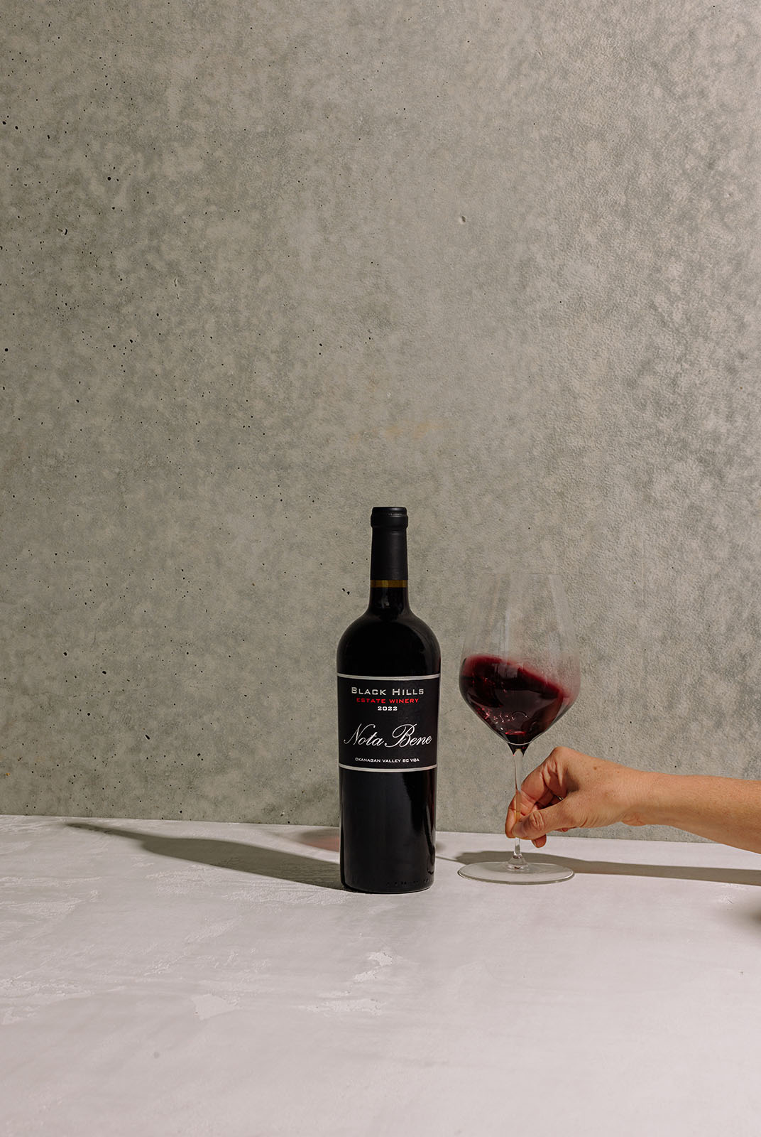A bottle of red wine labeled "Black Hills Nota Bene" is placed on a light gray surface against a textured gray background. A person's hand is holding a wine glass filled with red wine to the right of the bottle.