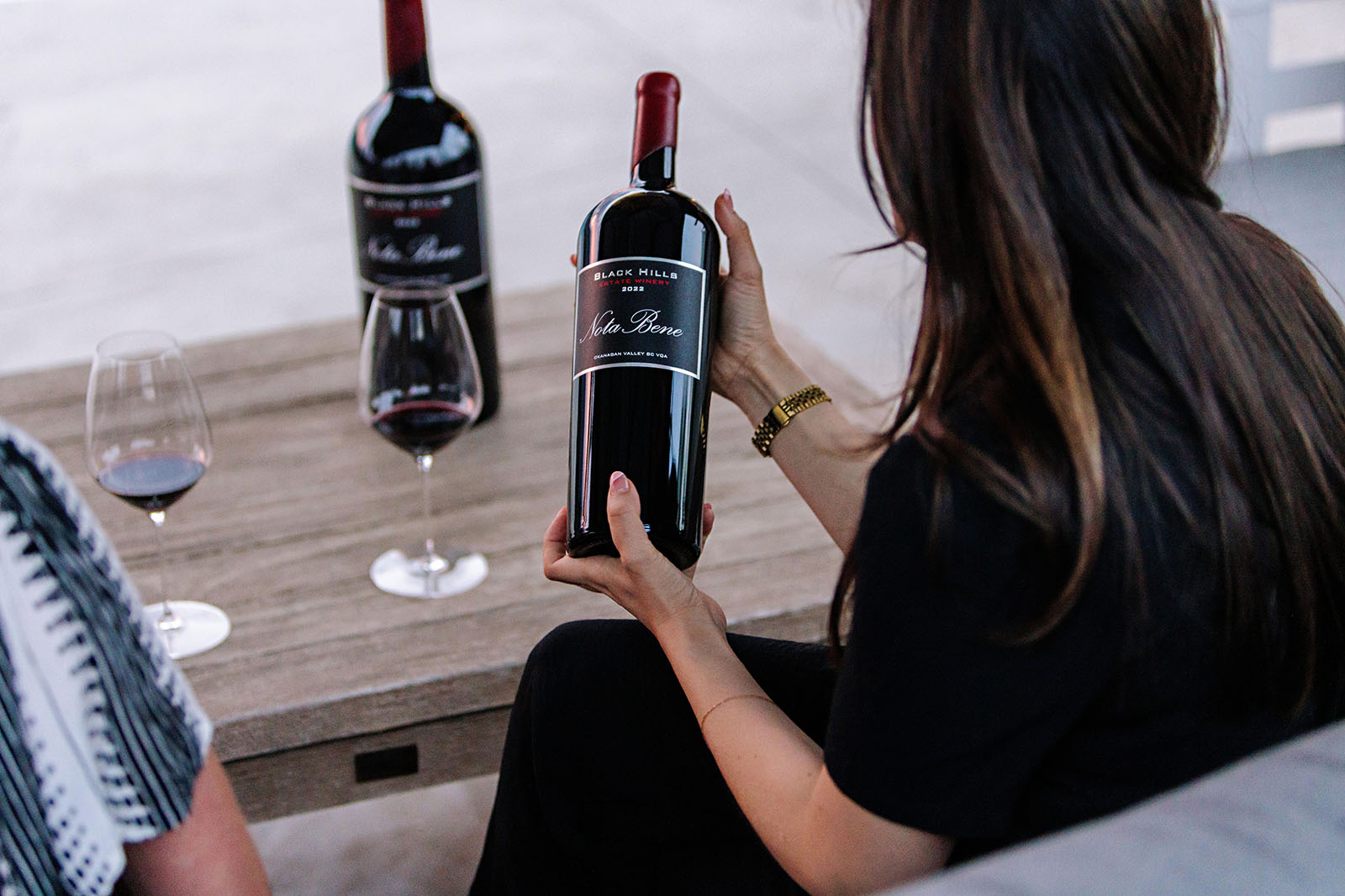 A person with long hair, dressed in black, is sitting at a wooden table holding a large bottle of red wine labeled "Black Hills." Another similar bottle and two glasses of red wine are on the table. The scene appears to be a relaxed setting.