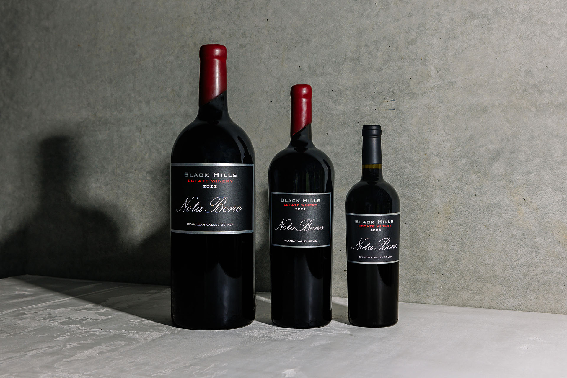 Three Black Hills Estate Winery "Nota Bene" wine bottles of different sizes are displayed against a gray background. The three sizes are large, medium, and small, each with a dark label featuring red and white text.